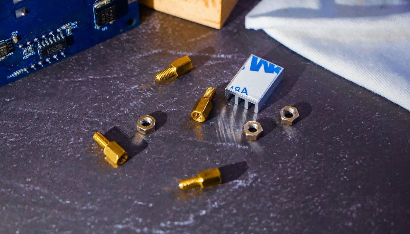 A small aluminium heatsink with adhesive thermal tape on its base, laid out beside brass standoffs and nuts next to a blue circuit board.